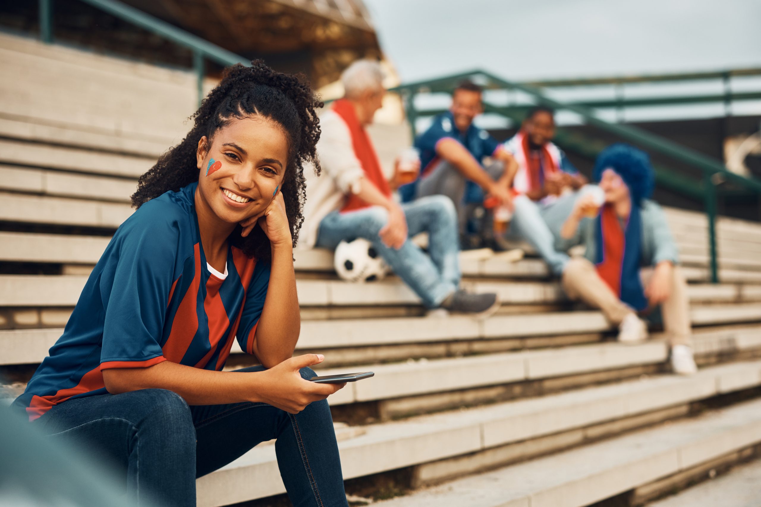 Happy African American woman in soccer jersey using smart phone and looking at camera.